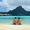 a honeymooning couple sits on the beach overlooking the ocean and resort bungalows